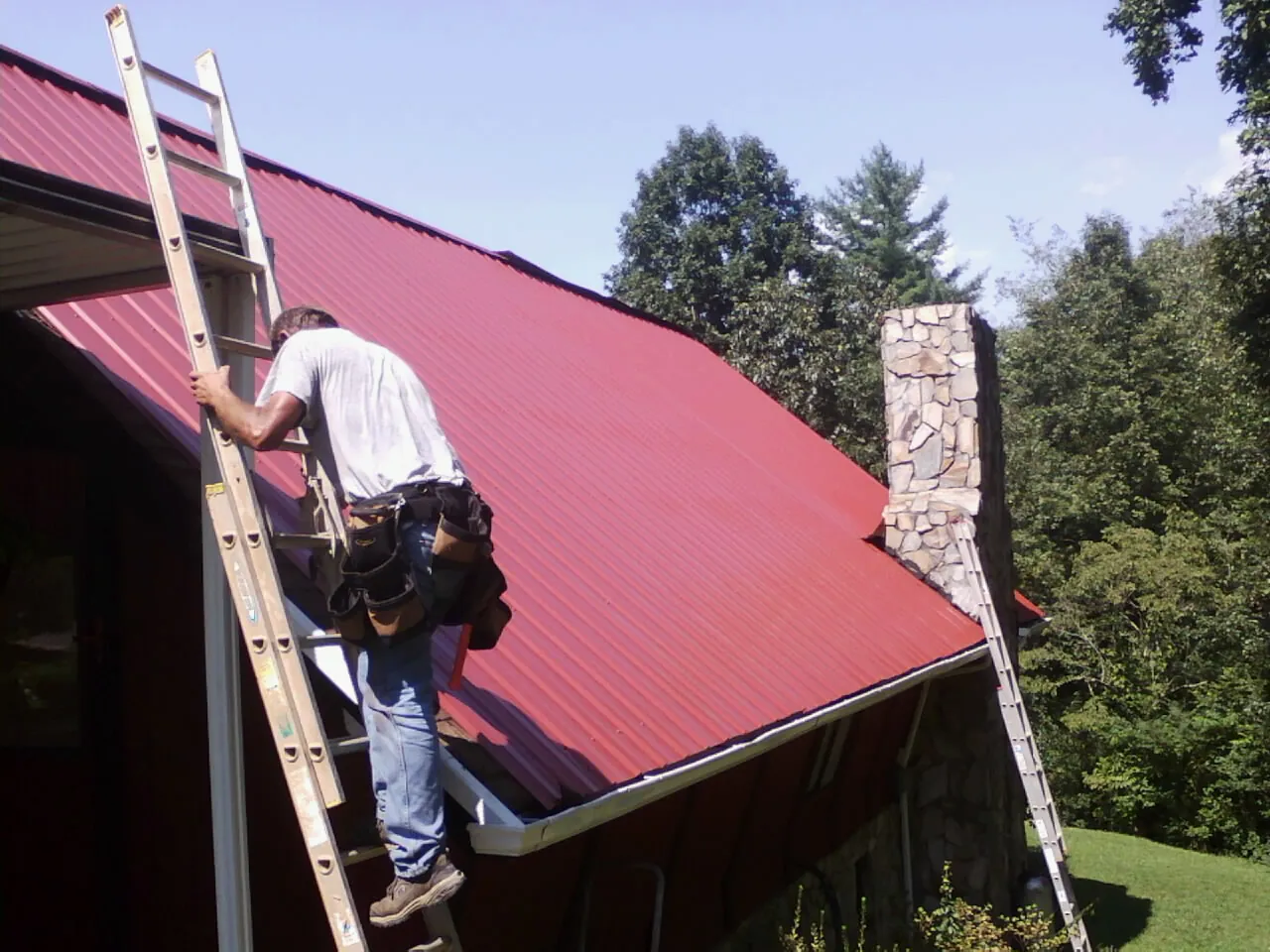Roofer on a ladder beside a red metal roof and stone chimney — Soffit & Fascia Repair by Roofing Brothers in Chicago Heights